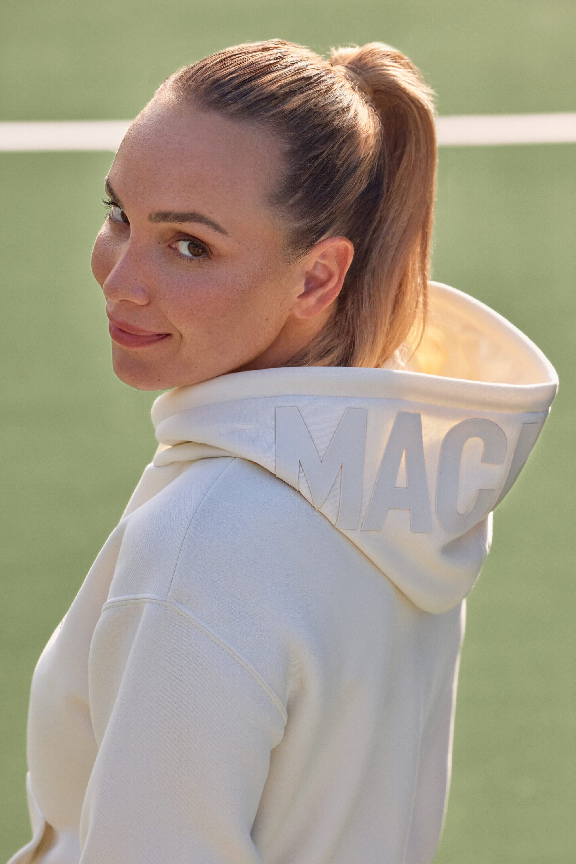 Donna Vekic wearing a cream hooded sweatshirt with branding on the hood, looking back over her shoulder outdoors.