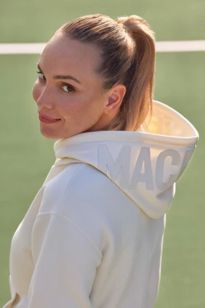 Donna Vekic wearing a cream hooded sweatshirt with branding on the hood, looking back over her shoulder outdoors.