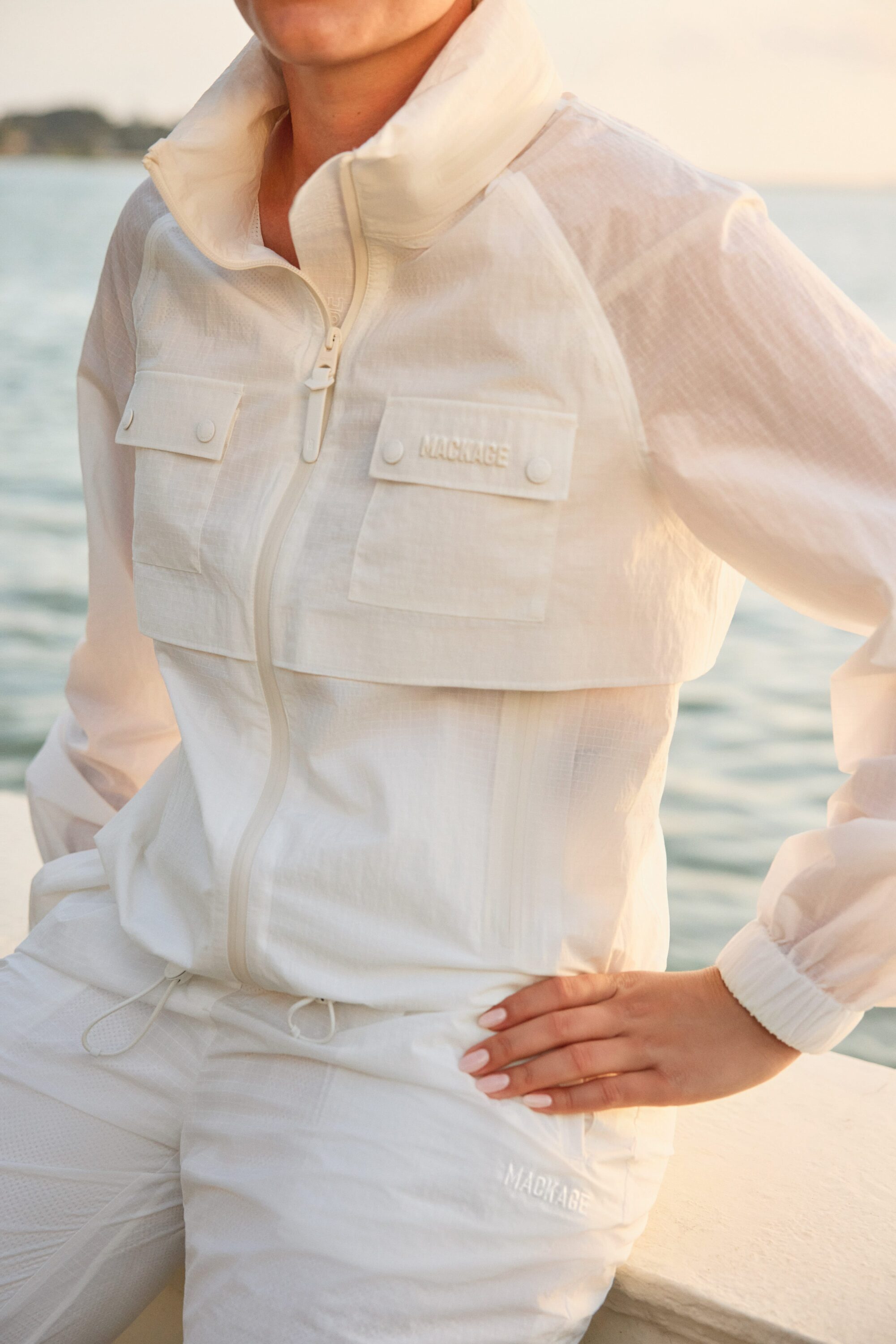 Model wearing a white zip-up windbreaker jacket with matching trousers outdoors near water