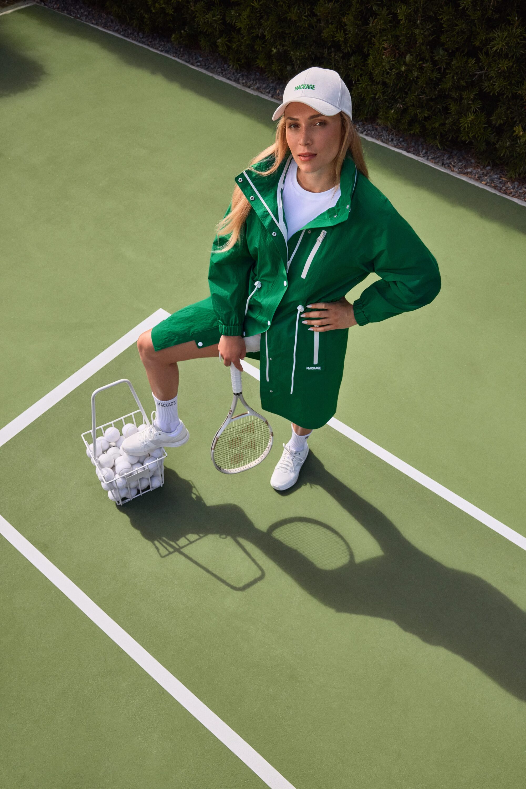 Donna Vekić wearing a green Mackage coat and white cap standing on a tennis court next to a basket of tennis balls