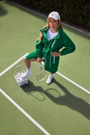 Donna Vekić wearing a green Mackage coat and white cap standing on a tennis court next to a basket of tennis balls