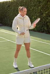 Donna Vekic holding a tennis racket on a court wearing a white puffer vest and shorts
