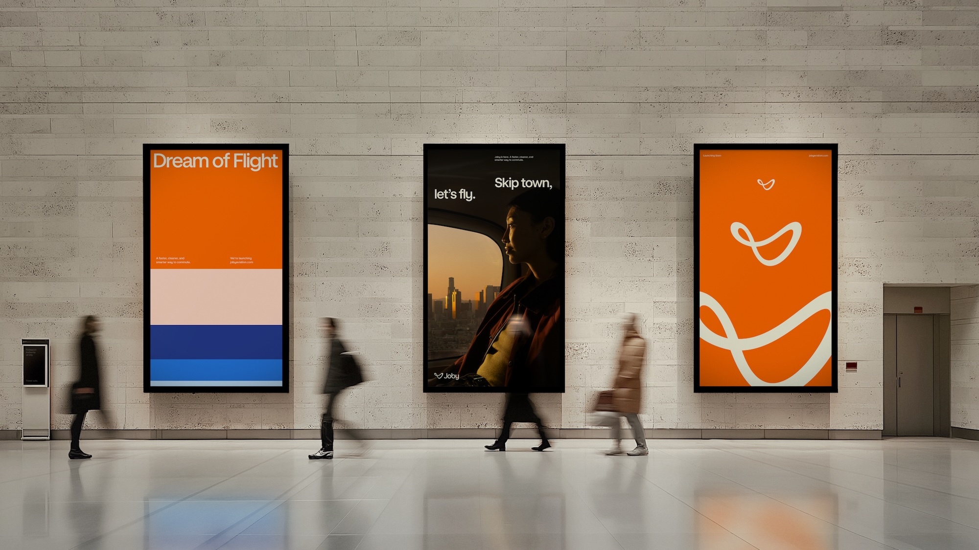 Three vertical digital advertising screens mounted on a stone tile wall in a modern public transit terminal, with blurred commuters walking past.