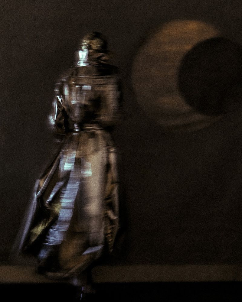 Person in a long, reflective coat walking past a wall with a circular light projection, creating a blurred and artistic effect.