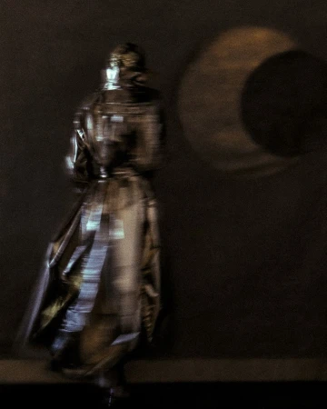 Person in a long, reflective coat walking past a wall with a circular light projection, creating a blurred and artistic effect.