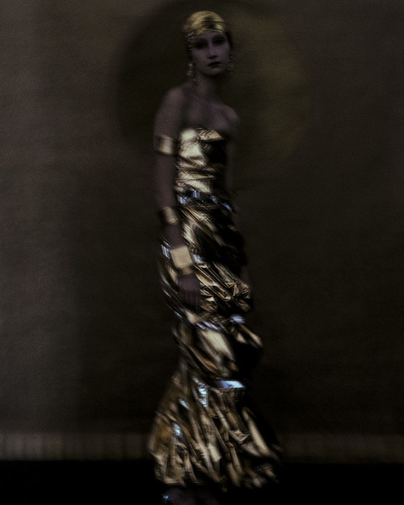 Elegant woman in golden strapless dress and gold headpiece stands against a neutral backdrop in a glamorous fashion portrait.