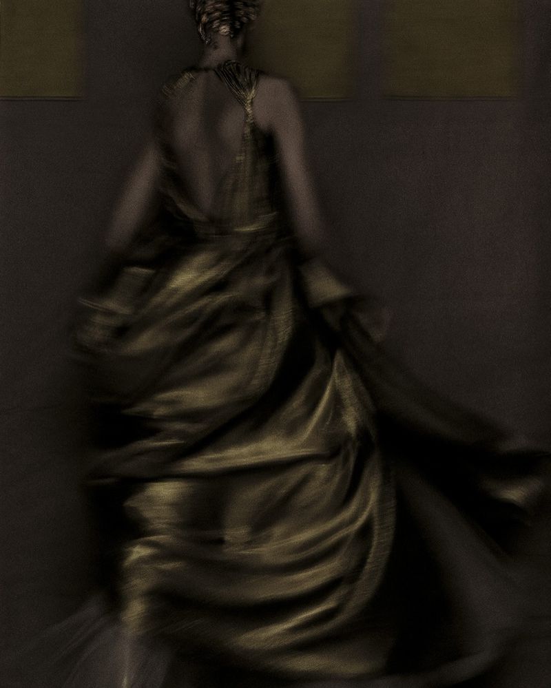 Back view of an adult woman in motion, wearing a flowing gold dress, in a dimly lit interior with diffused lighting and soft focus.