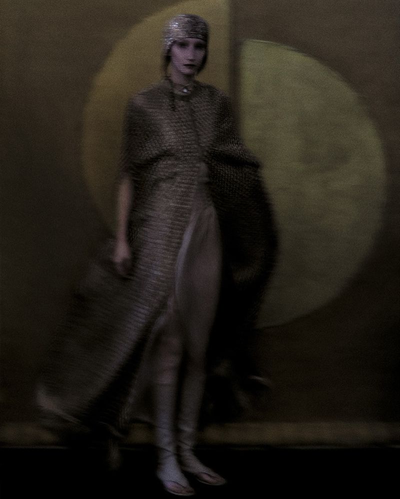 A woman in a beaded head covering and gown poses in front of a mottled brown and tan backdrop for a fashion or editorial photograph.