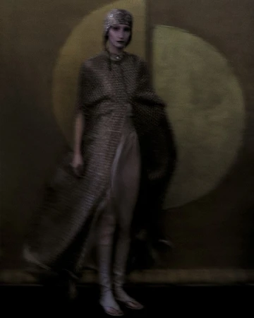 A woman in a beaded head covering and gown poses in front of a mottled brown and tan backdrop for a fashion or editorial photograph.