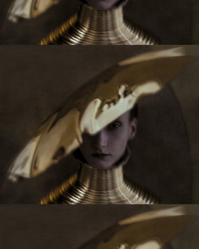 Woman in avant-garde fashion featuring a shiny gold hat and a layered metallic collar. Studio lighting highlights the sculptural design.
