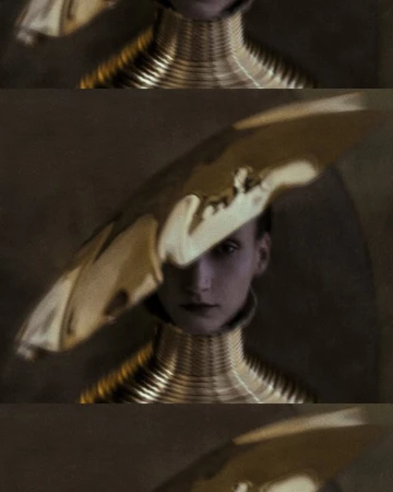 Woman in avant-garde fashion featuring a shiny gold hat and a layered metallic collar. Studio lighting highlights the sculptural design.