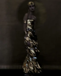 Model in shiny gold strapless dress and gold headband standing against dark background