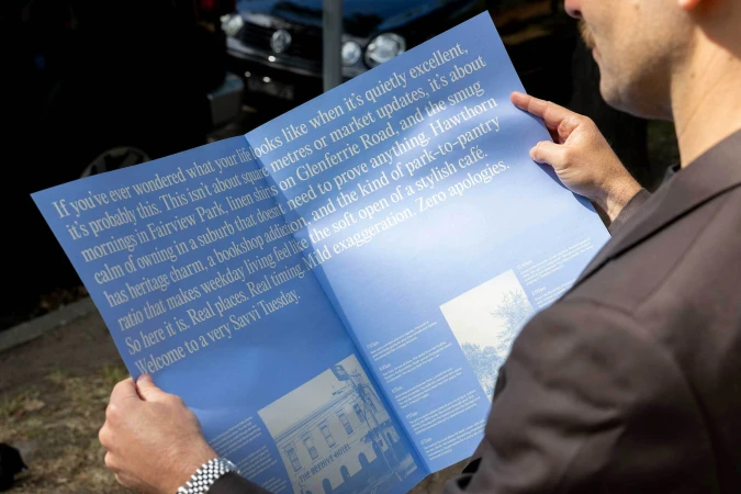 Man wearing a dark suit jacket holding an open blue brochure with white printed text outdoors.