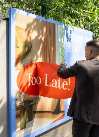 A man in a dark suit peeling an orange sticker off a large outdoor blue advertising billboard for Savvi Real Estate.