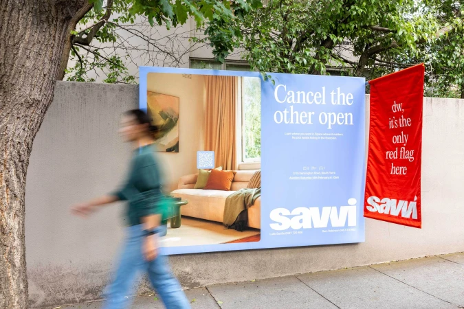 Outdoor advertising billboard for Savvi Real Estate on a concrete wall with a blurred pedestrian walking past