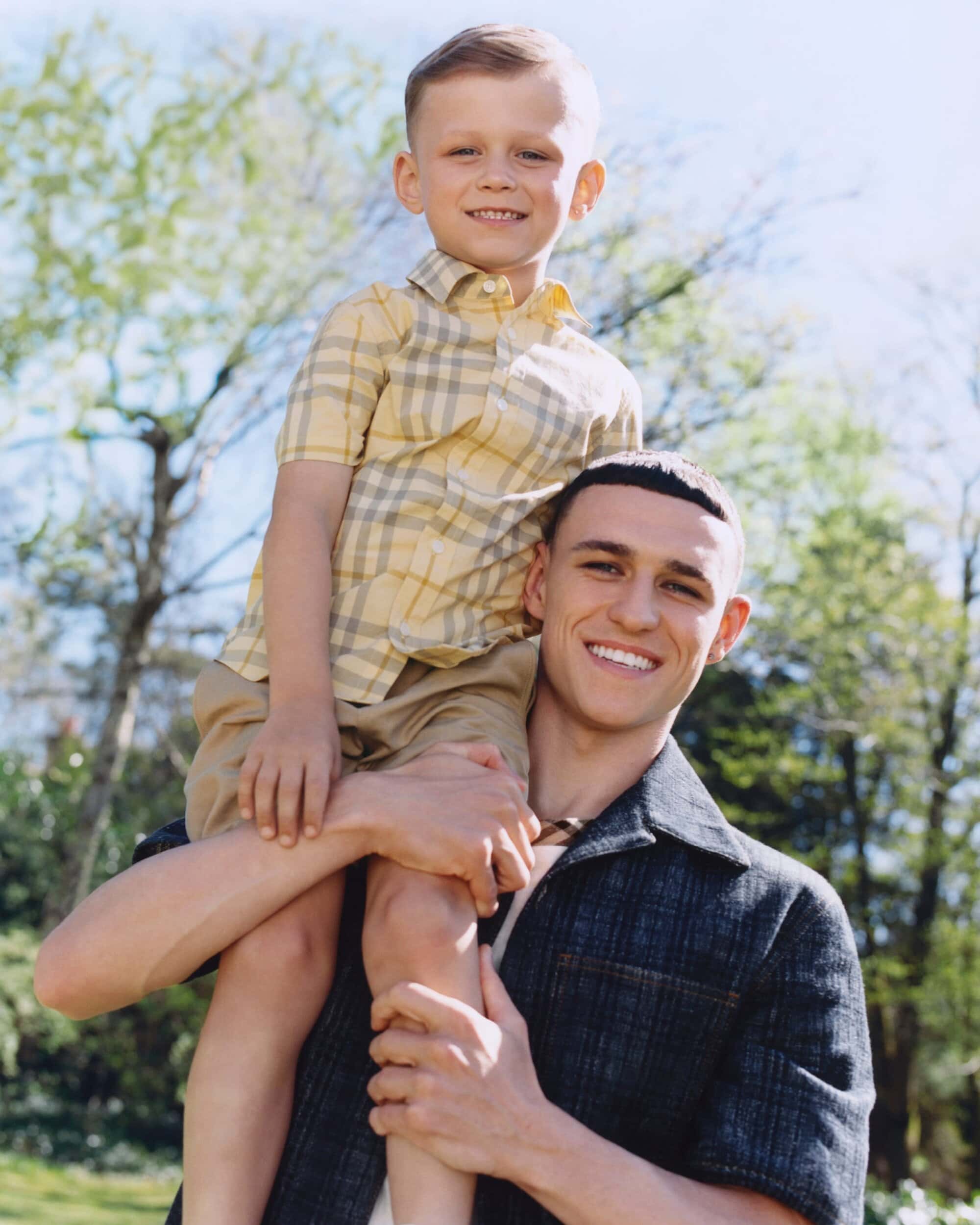 Phil Foden holding a child on his shoulders outdoors wearing a dark patterned shirt against trees