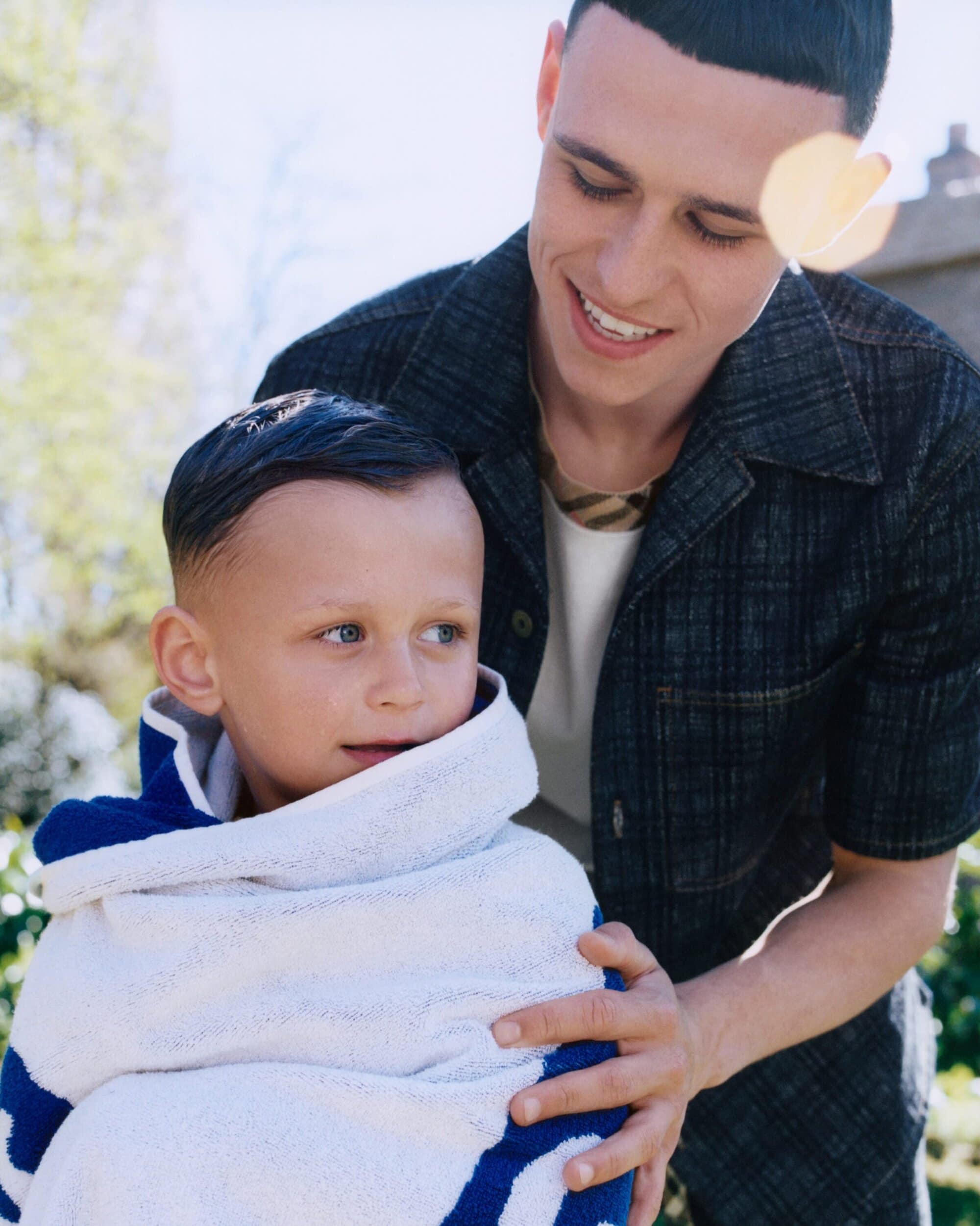 Phil Foden smiling while wrapping a young child in a blue and white towel outdoors