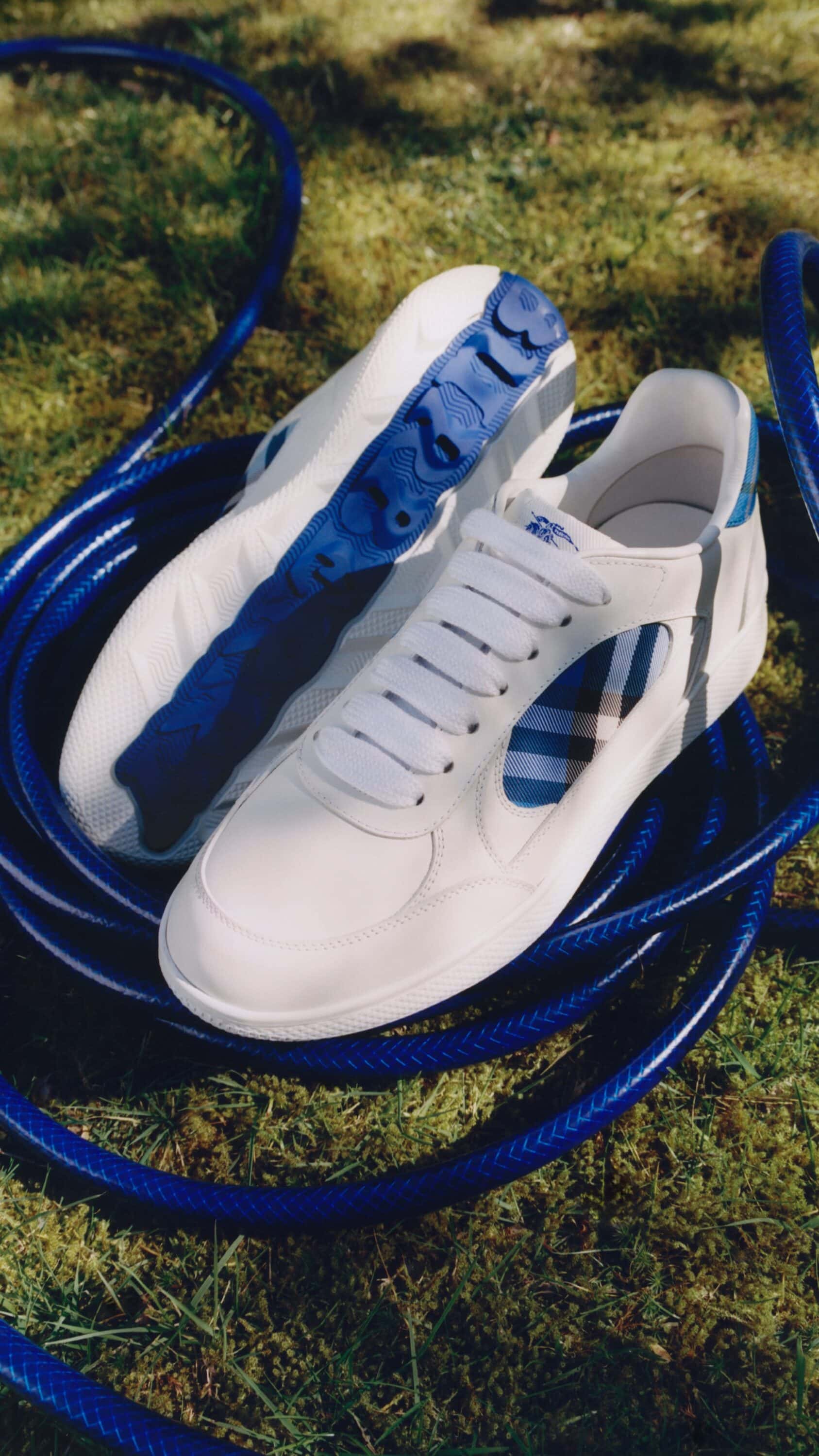 White leather sneakers with blue plaid accents resting on a coiled blue hose over a grassy lawn background.