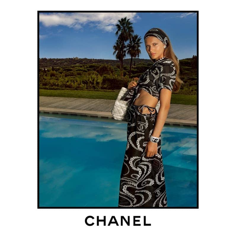 Model wearing a printed black and white outfit beside a swimming pool under a bright sky with palm trees.