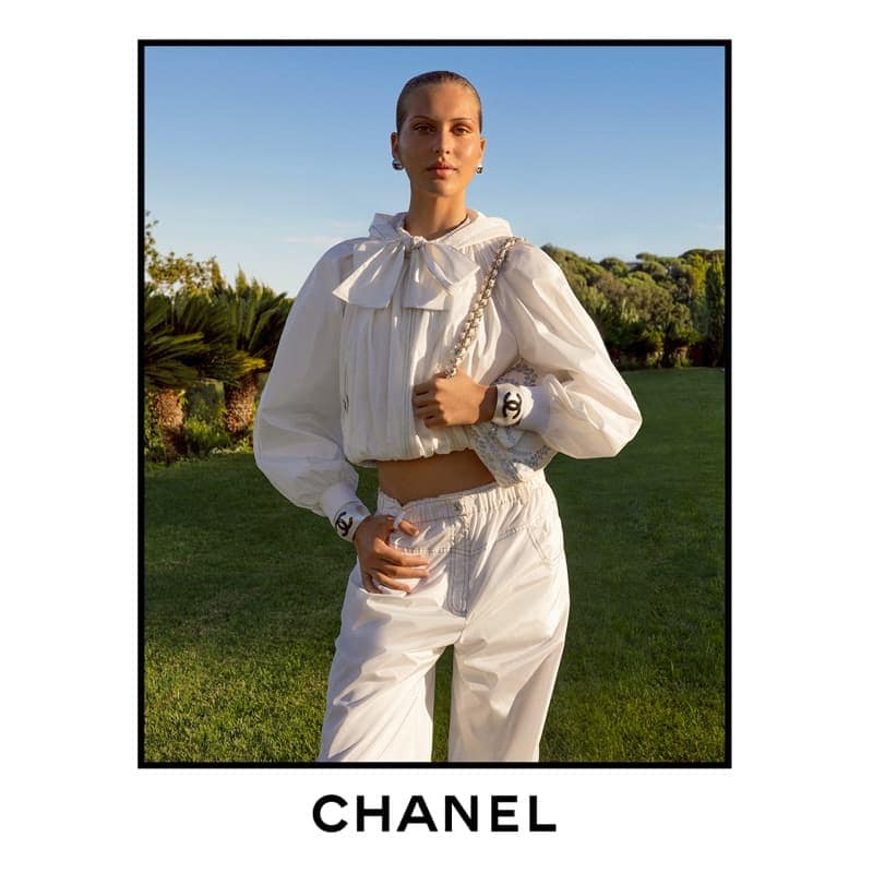 Model in a white blouse and matching trousers standing outdoors on a green lawn against a blue sky