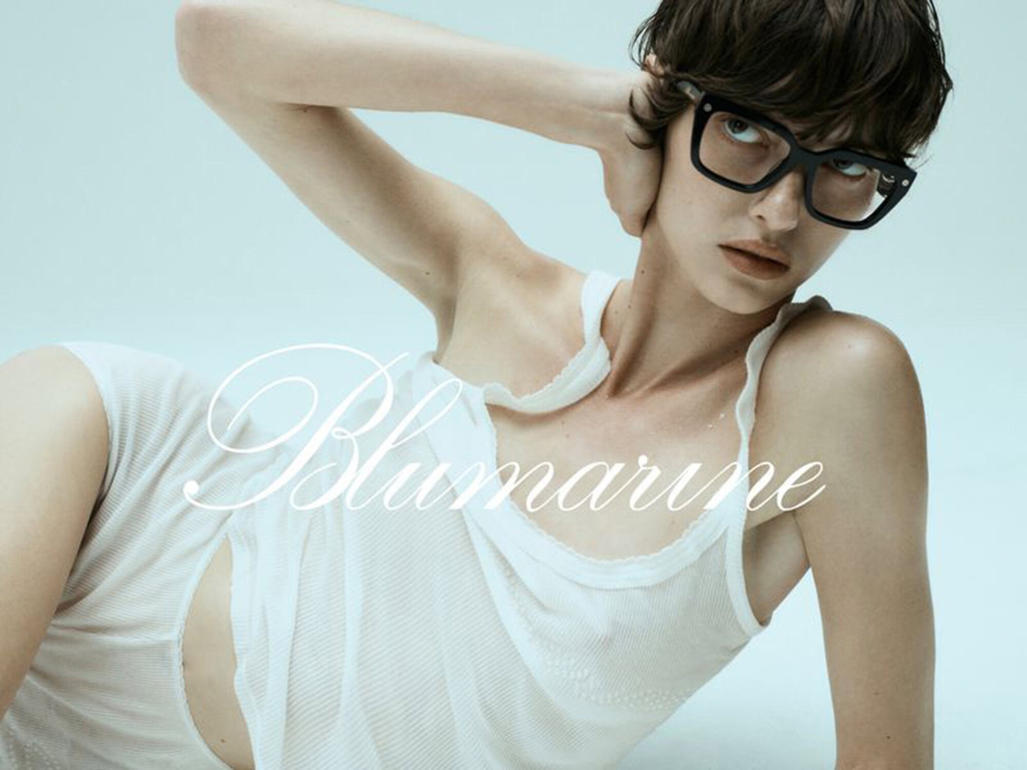 Model with short dark hair wearing thick black eyeglasses and white top against a pale blue background for Blumarine.