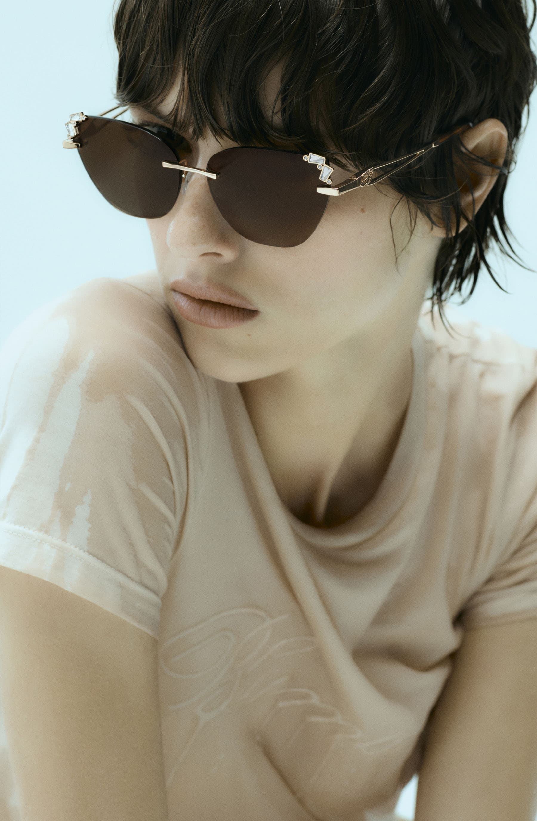 Model wearing dark tinted sunglasses and a light-coloured top against a pale blue background for Blumarine Eyewear 2025.