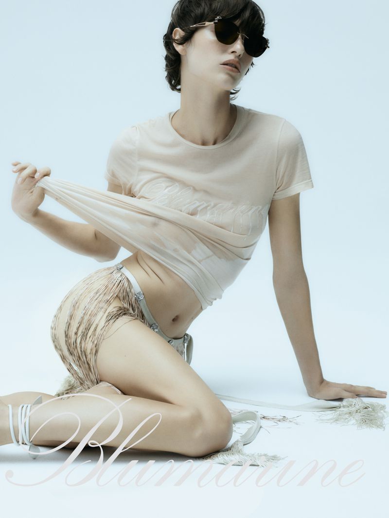 Model wearing dark sunglasses and a sheer top sitting against a pale blue seamless background