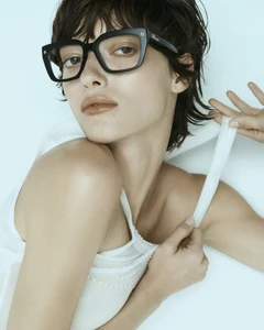 Model wearing thick black frame glasses and a white top against a pale background for Blumarine Eyewear 2025 campaign.