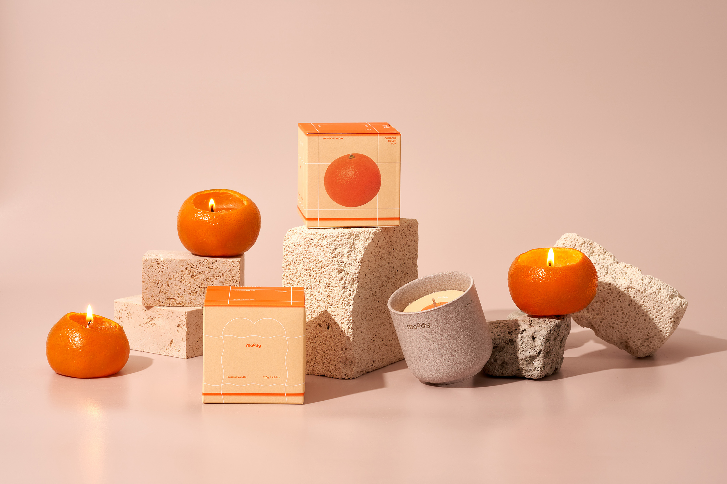 Orange scented candles and matching packaging boxes arranged on rough stone blocks against a neutral pinkish studio background.