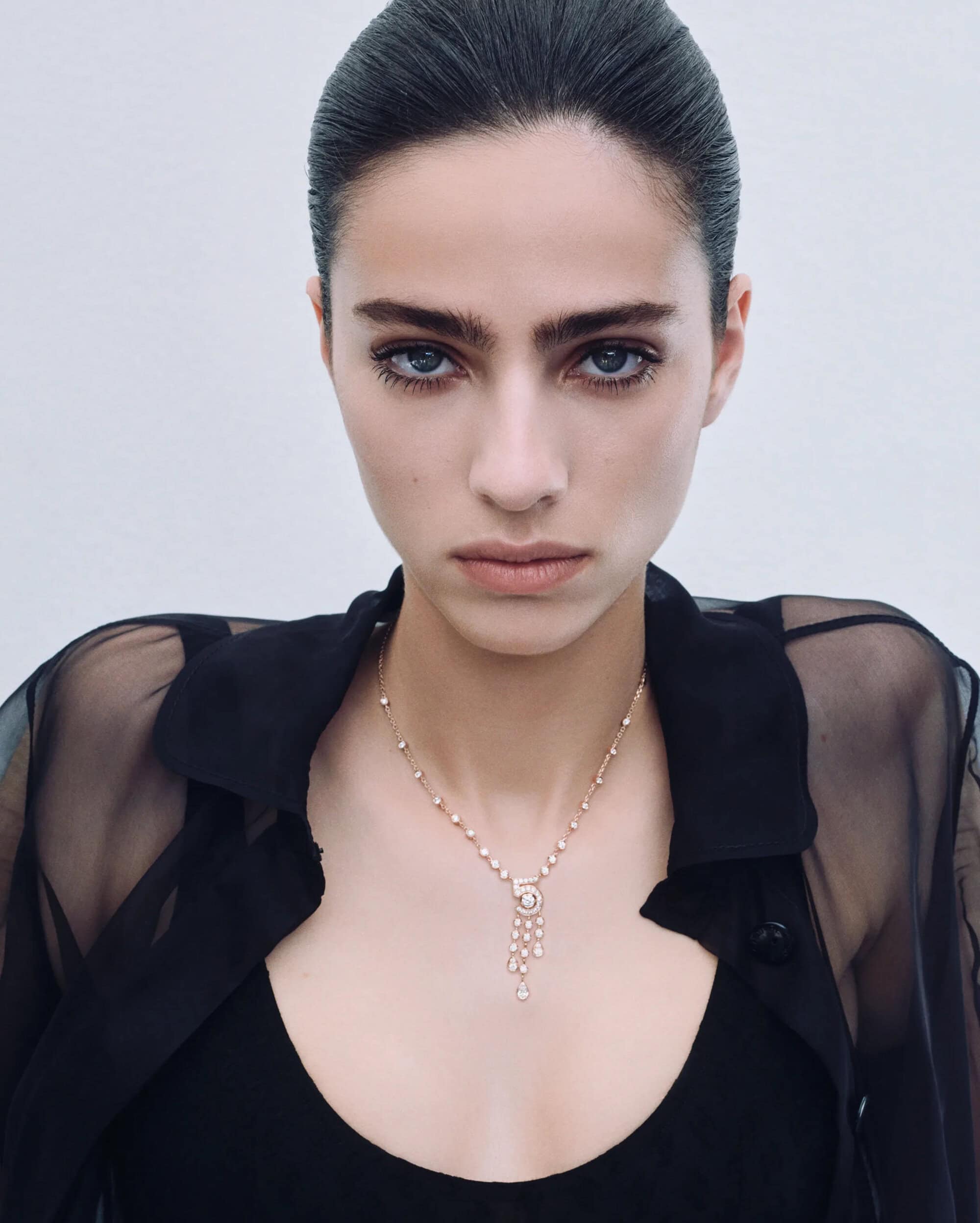 Close-up portrait of a model wearing a fine jewelry necklace and black sheer top against a white background.