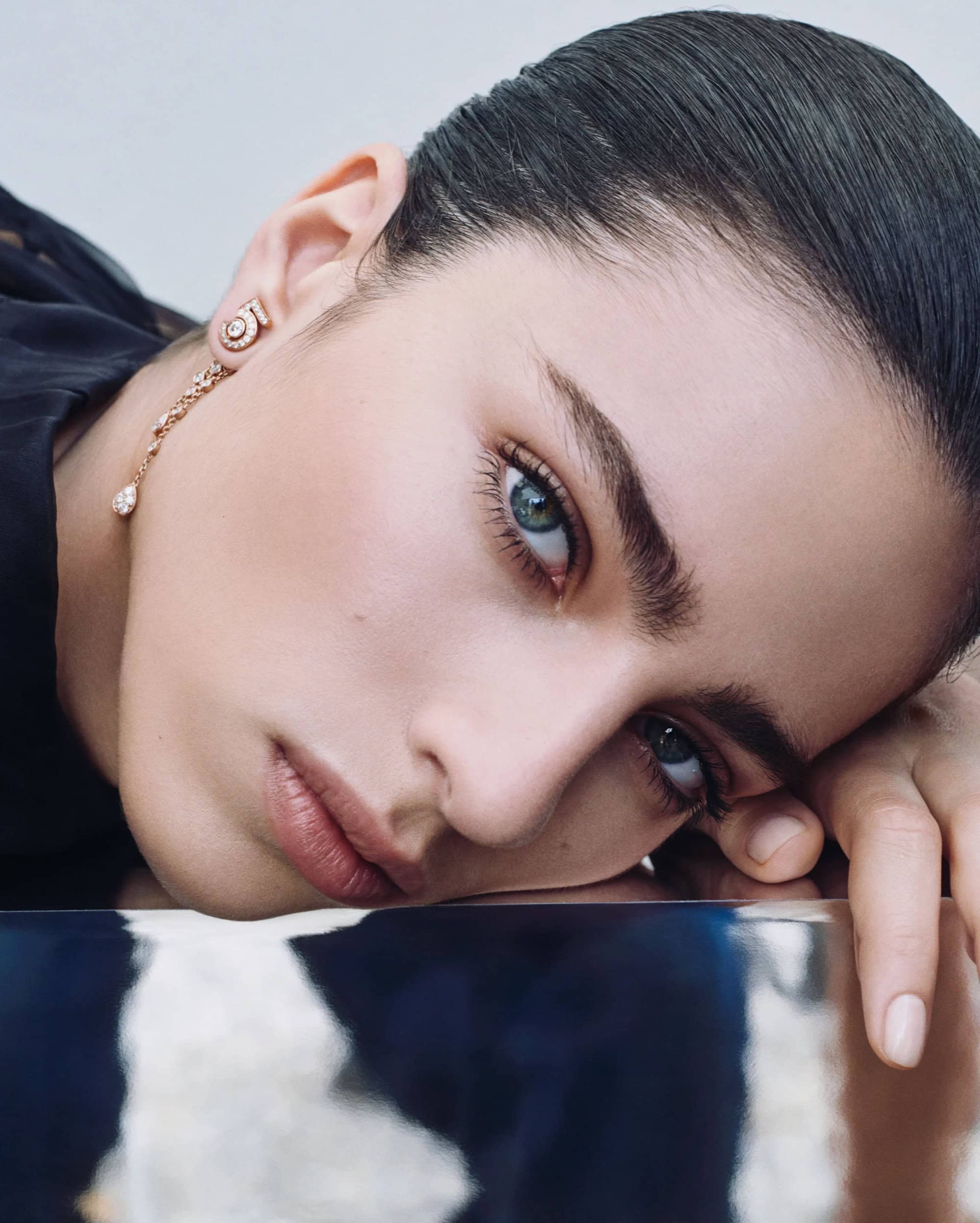 Close-up portrait of a female model wearing drop diamond earrings against a reflective metallic surface for Chanel fine jewelry.
