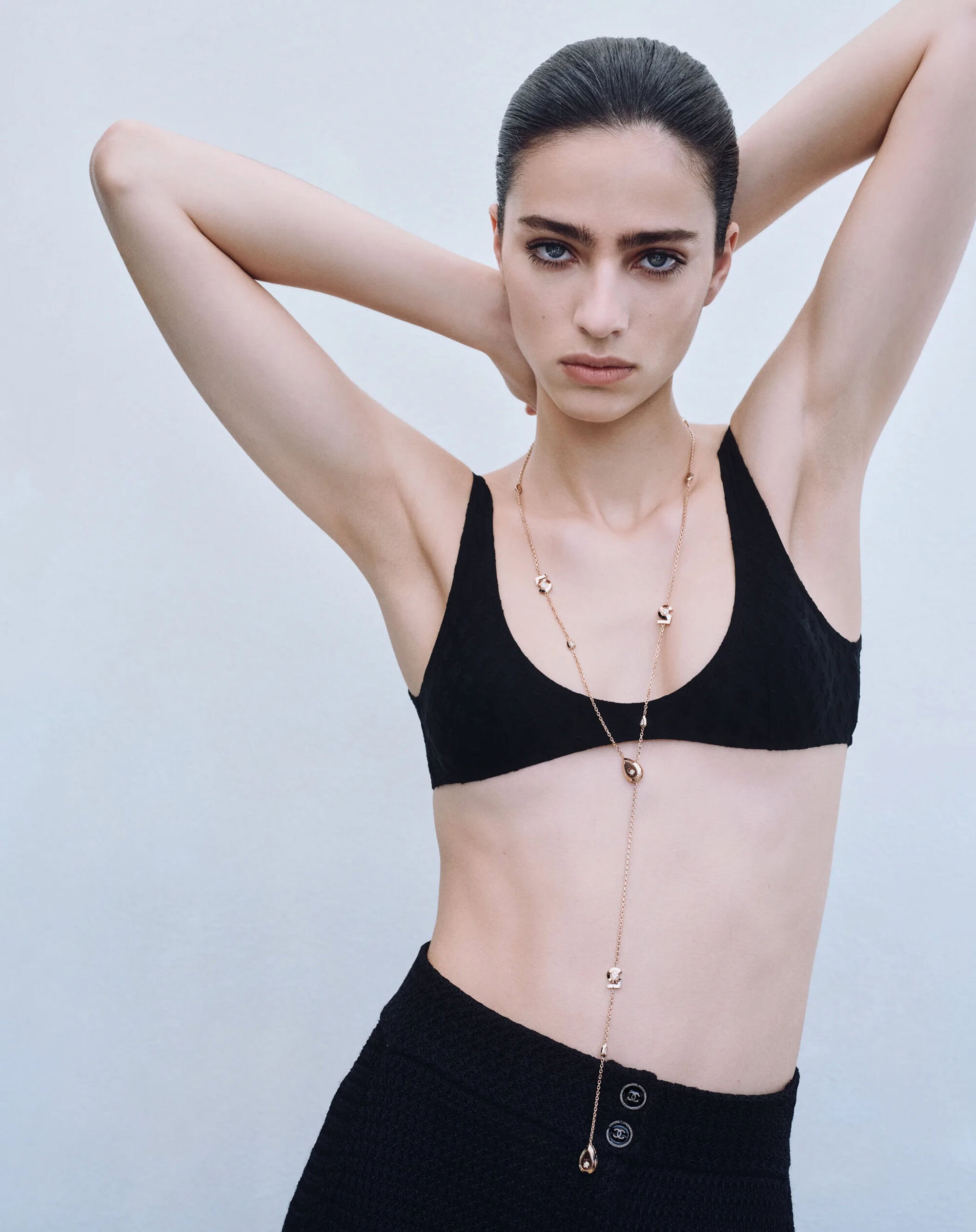 Model posing with arms raised wearing a black top and long gold pendant necklace against a light grey background