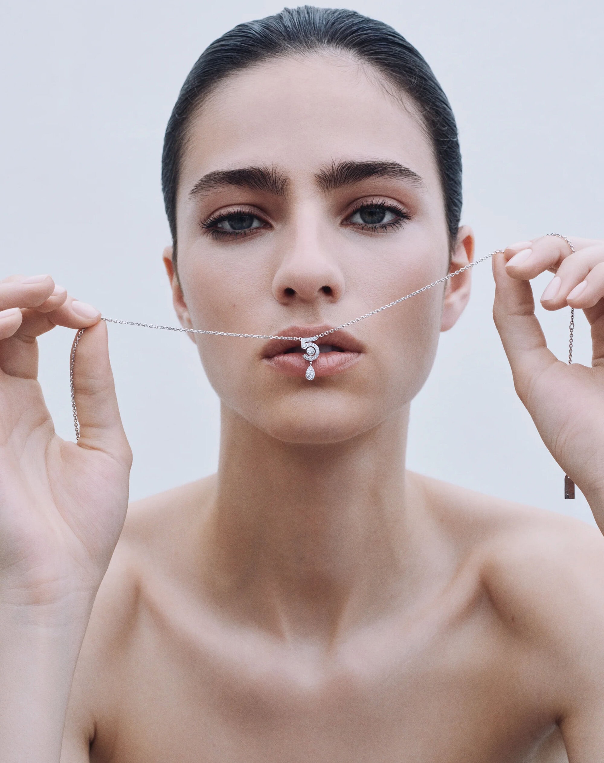 Close-up portrait of a model holding a delicate silver necklace with a diamond pendant near lips against a light background.