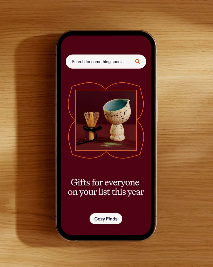 Smartphone displaying Etsy mobile app interface resting on a light wooden surface with a soft shadow cast alongside it.