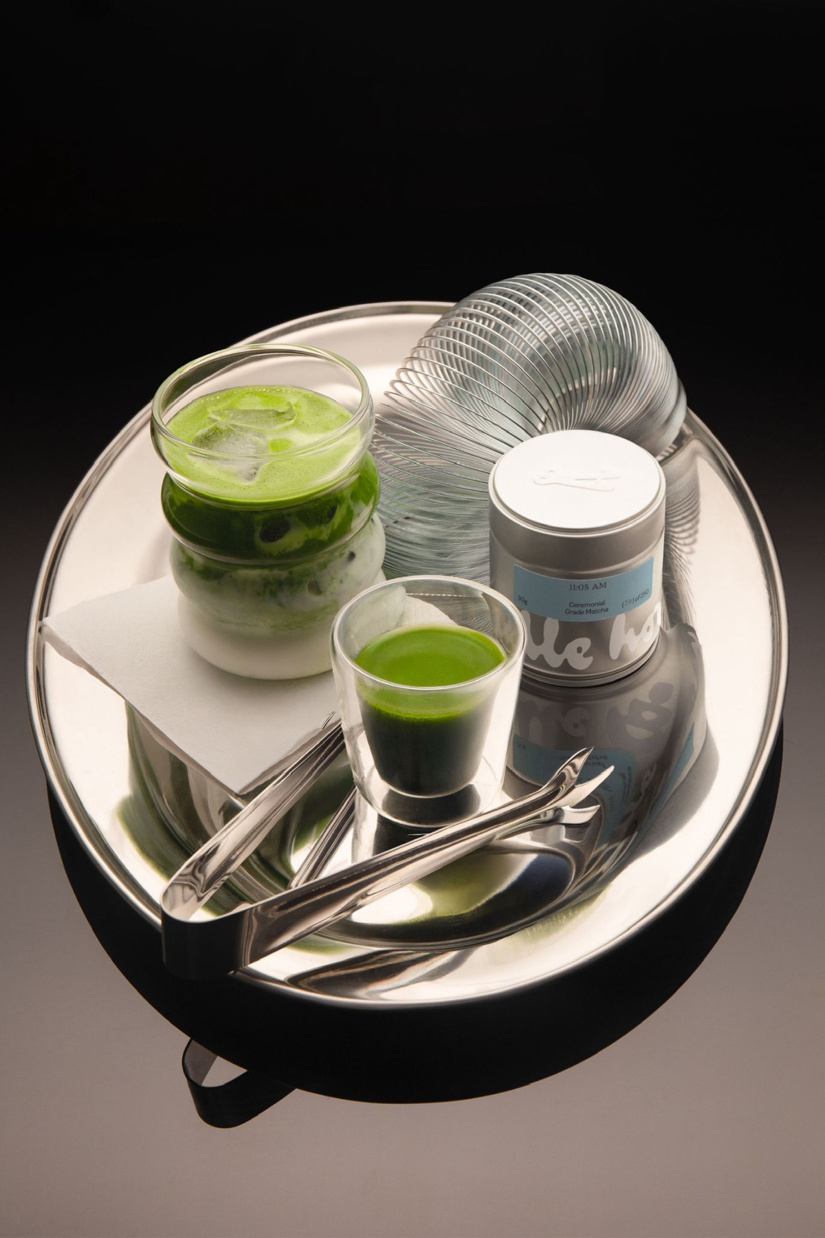 Green iced matcha drink, small green shot glass, silver tin, and metal slinky on a shiny silver oval tray