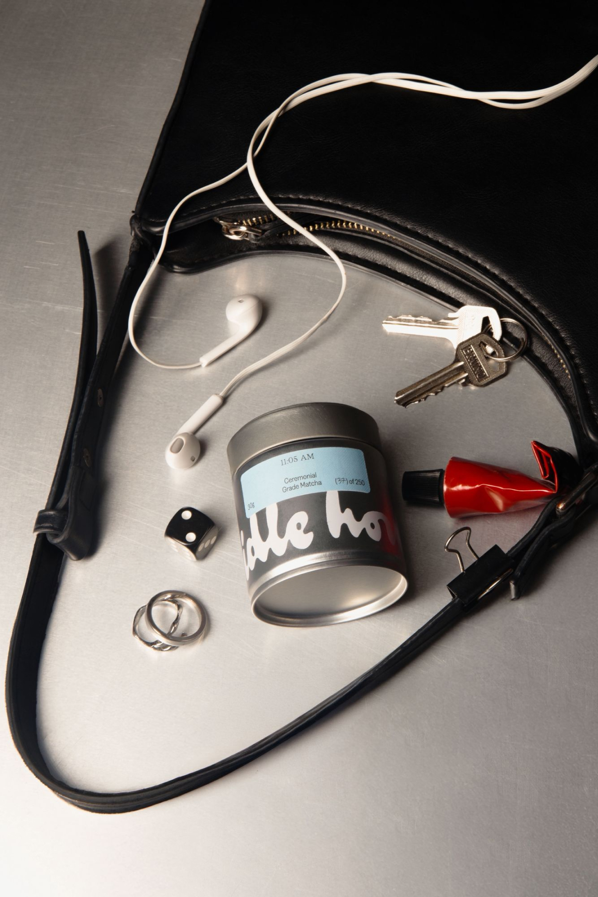 An open black handbag on a metallic surface containing keys, white earphones, a tin container, dice, and rings.