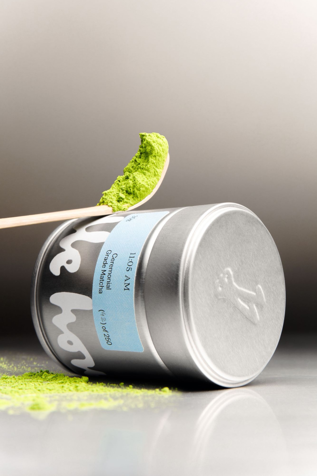 Silver matcha tea tin resting horizontally on a gradient surface with a wooden spoon holding a scoop of bright green matcha powder