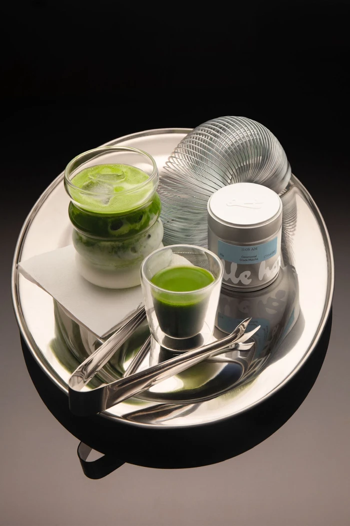 Green iced matcha drink, small green shot glass, silver tin, and metal slinky on a shiny silver oval tray