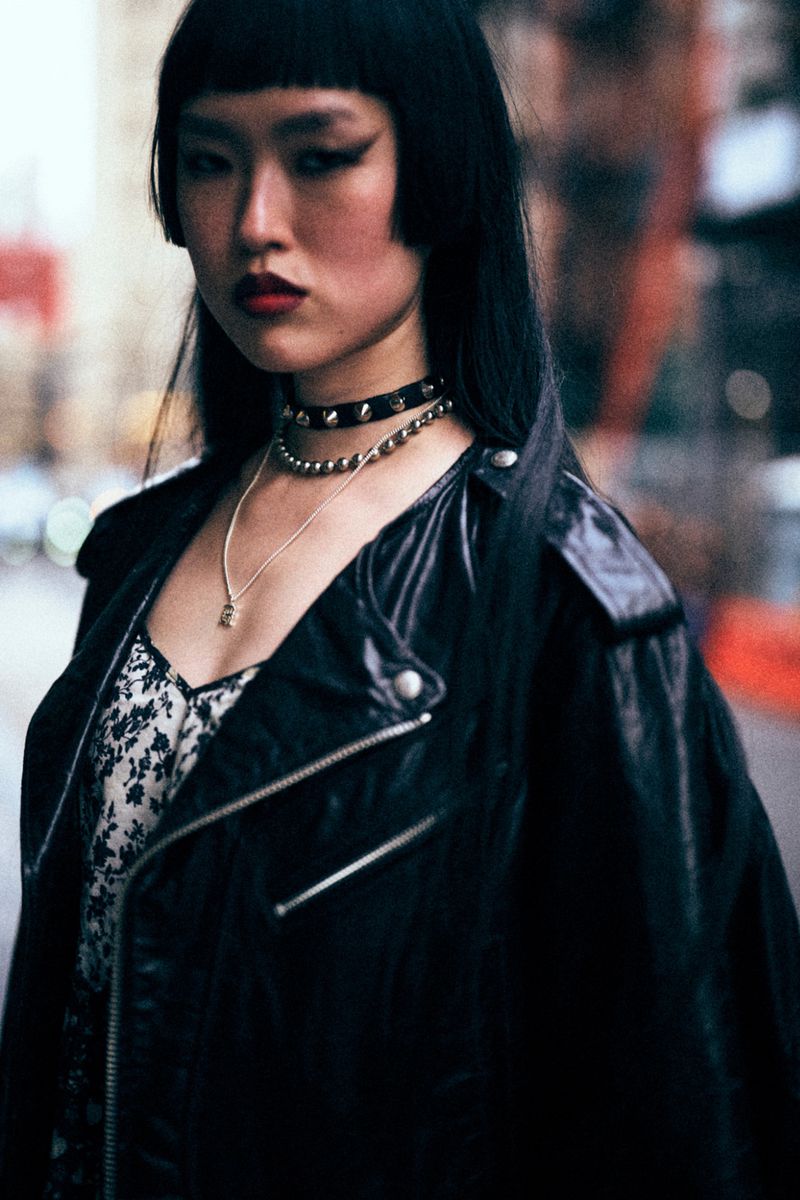 Close-up of an Asian woman with dark hair and heavy eye makeup wearing a black leather jacket against an urban backdrop.