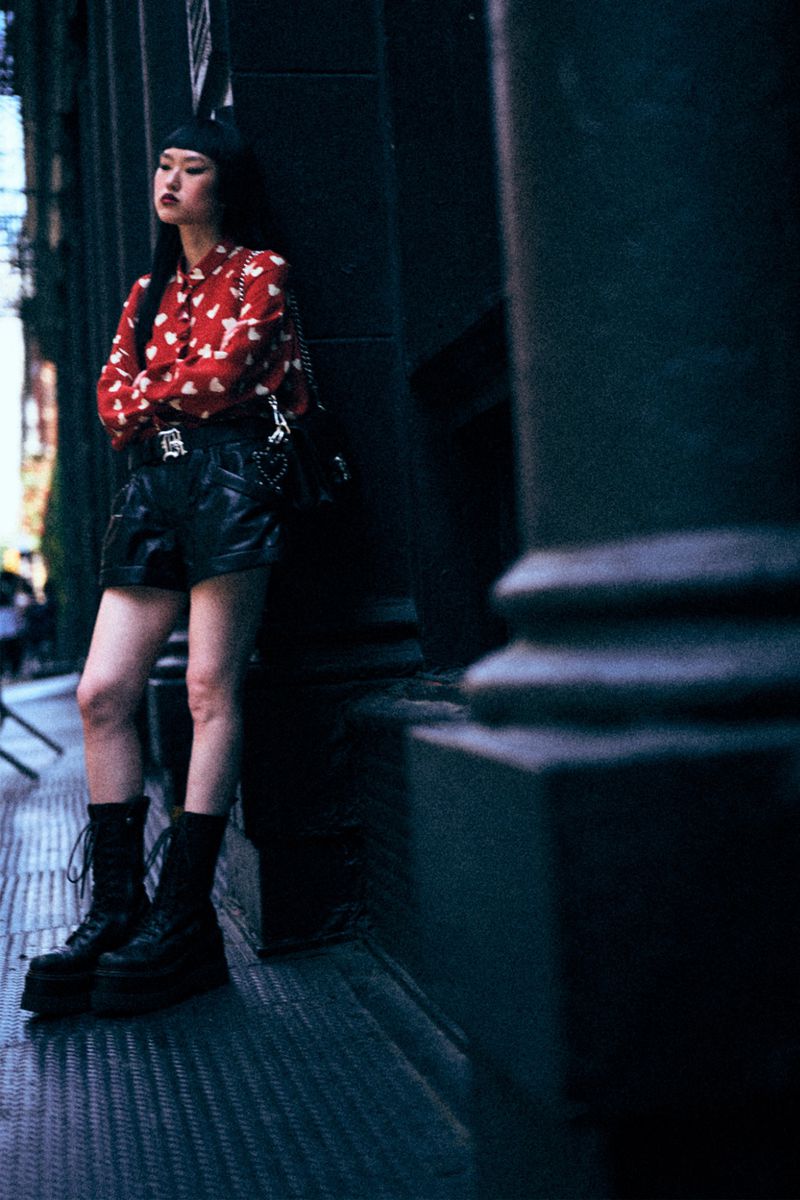 A young adult female stands leaning against a dark stone column in an urban setting wearing a red patterned blouse and black shorts and boots.