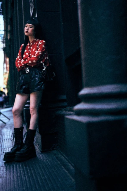 A young adult female stands leaning against a dark stone column in an urban setting wearing a red patterned blouse and black shorts and boots.