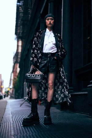 Stylish Asian woman in black leather skirt, platform boots, floral coat, beanie, and bag with silver rings in a dark urban setting.