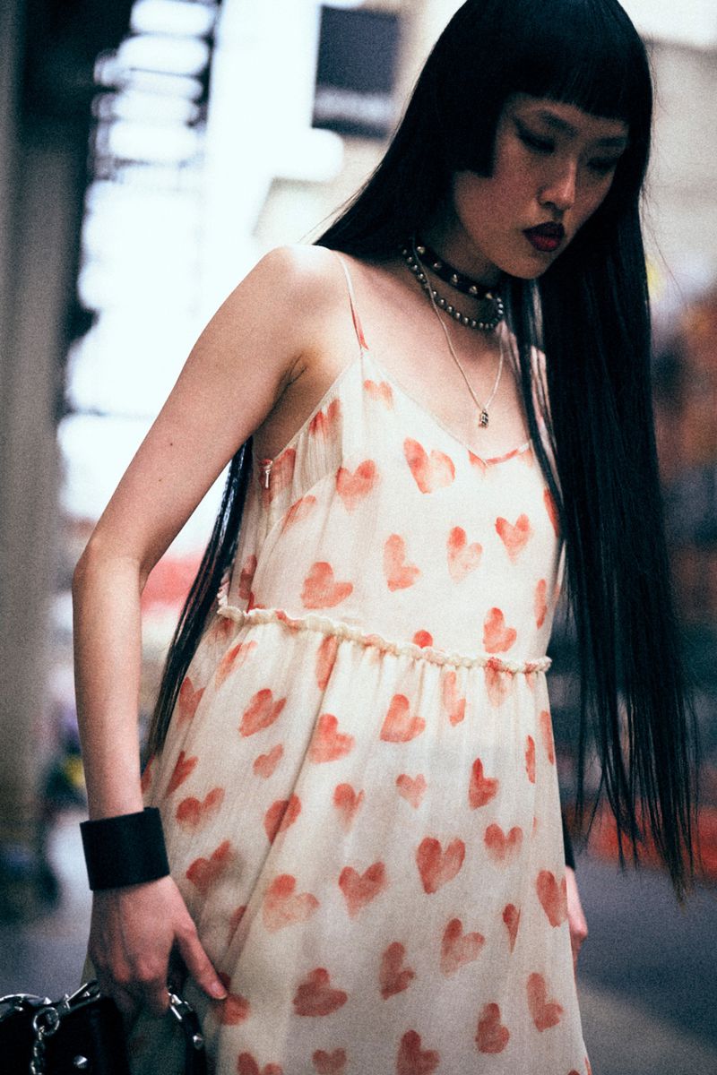An Asian woman wearing a white dress with red hearts poses outdoors. She has long black hair, a choker, and is carrying a bag.