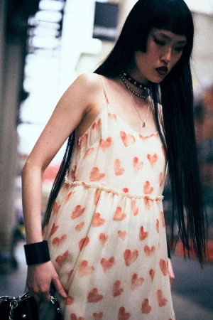 An Asian woman wearing a white dress with red hearts poses outdoors. She has long black hair, a choker, and is carrying a bag.