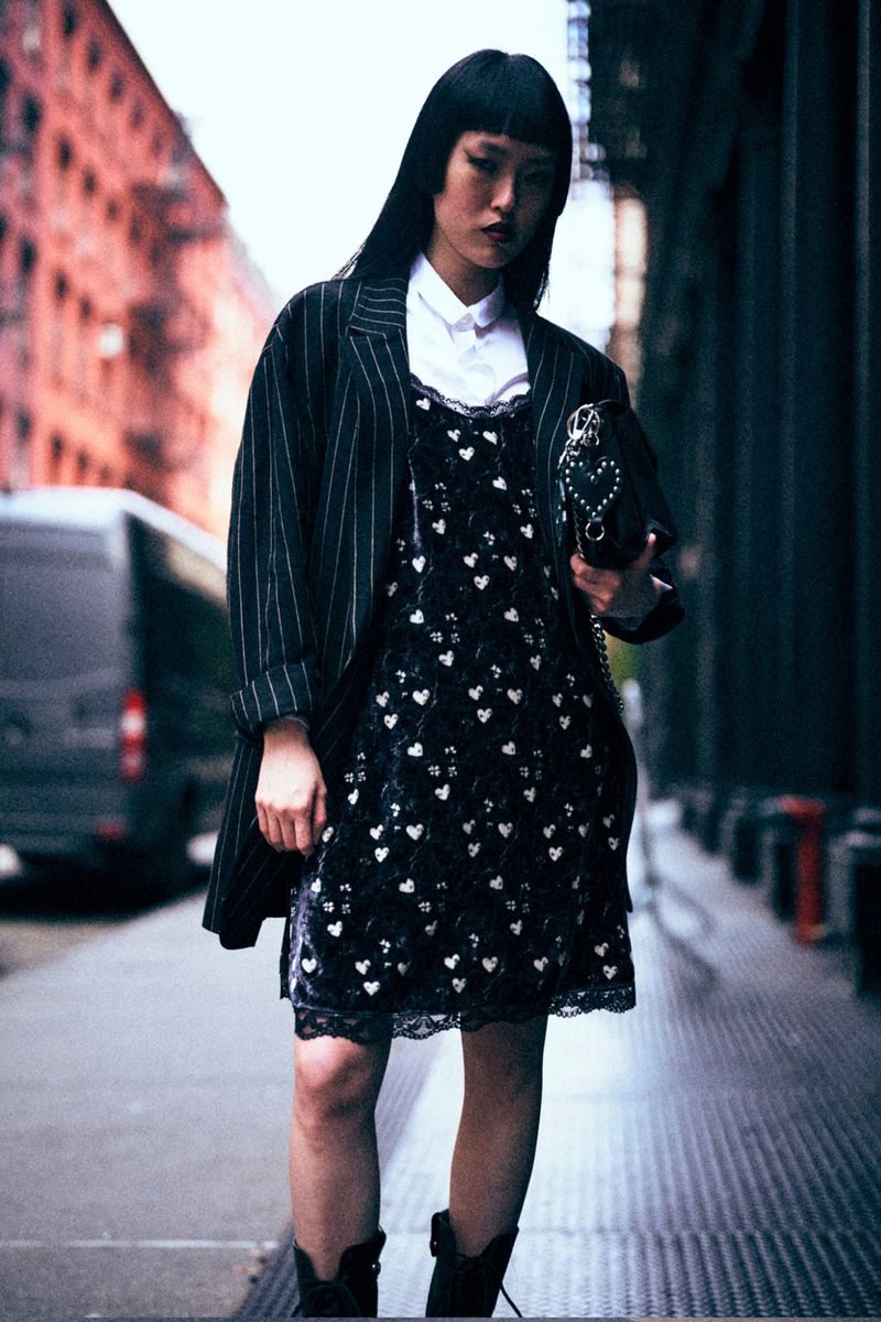 A stylish woman in a heart-printed dress, pinstripe jacket, and boots stands in an urban setting with an edgy, fashionable look.