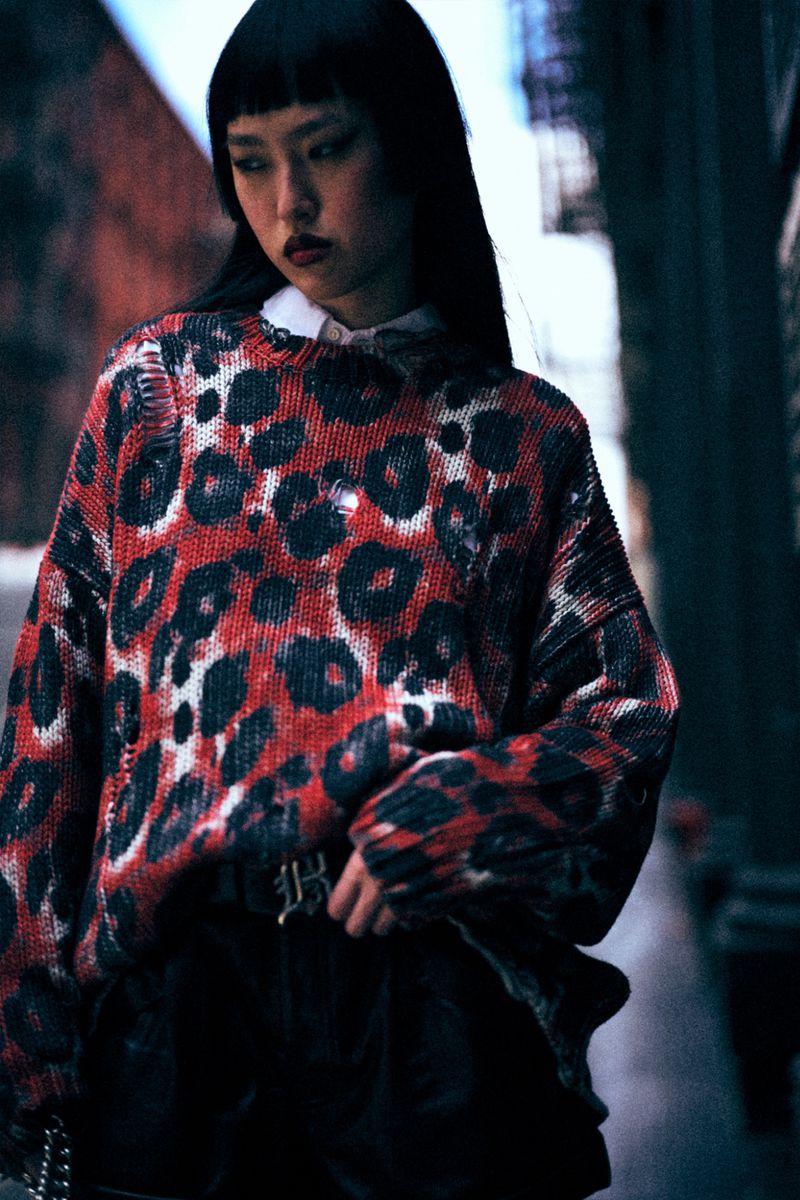Asian woman in a fashion shot, wearing a red and black animal print sweater, black bottoms, and a white collared shirt.