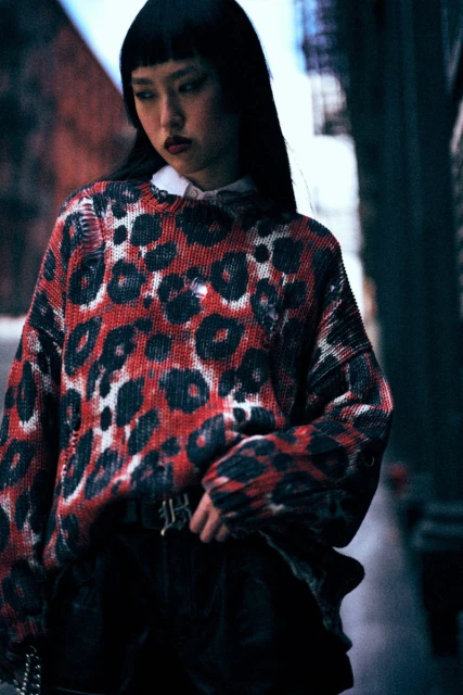 Asian woman in a fashion shot, wearing a red and black animal print sweater, black bottoms, and a white collared shirt.