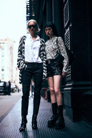 Two women pose on a city street; one with platinum blonde hair wears sunglasses, zebra-striped cardigan, and the other with bangs in floral blouse.