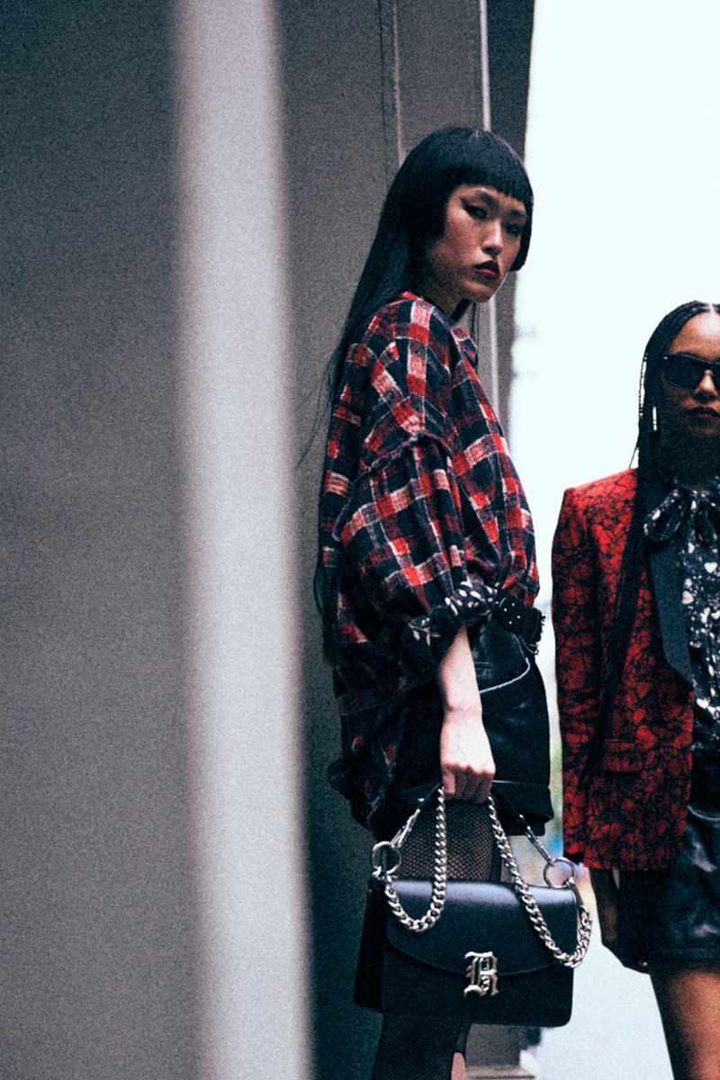 Two women pose outdoors wearing fashion apparel. One holds a black purse with a chain. The background features a concrete wall.