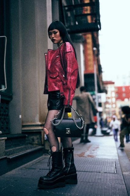 A woman poses in a red leather jacket, black skirt, and platform boots on a city sidewalk with buildings and people in the background.